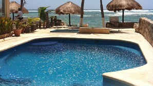 A pool area with a clear view of the ocean, surrounded by palm trees and thatched umbrellas.