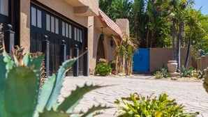 Uma passarela pavimentada conduz a um edifício com porta de garagem escura, cercado por cactos e plantas tropicais.