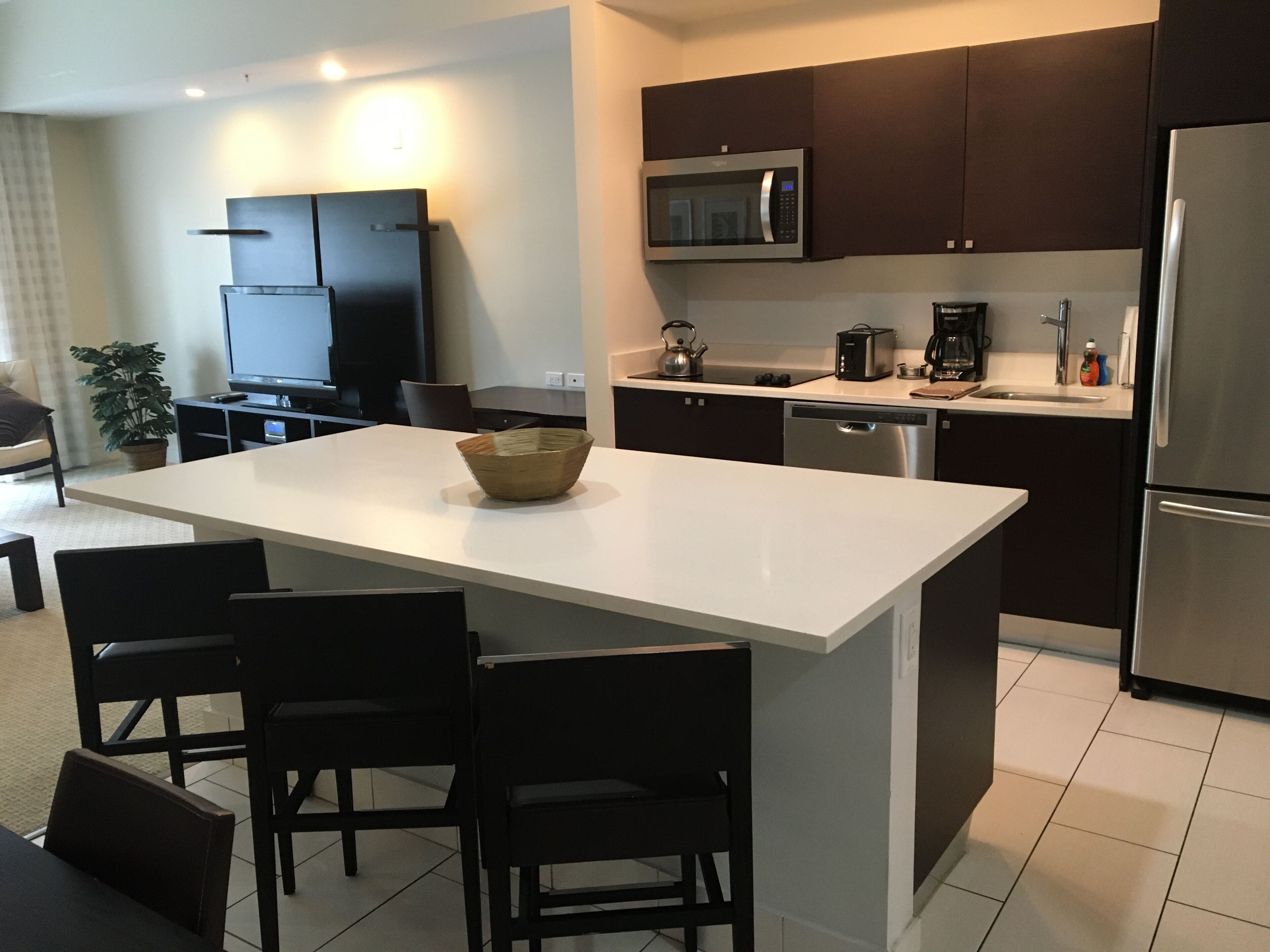 A modern kitchen with a central island, dark cabinetry, stainless steel appliances, and a flat-screen TV.