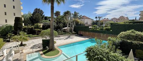 A swimming pool surrounded by greenery, with a palm tree and a hedge in the foreground.