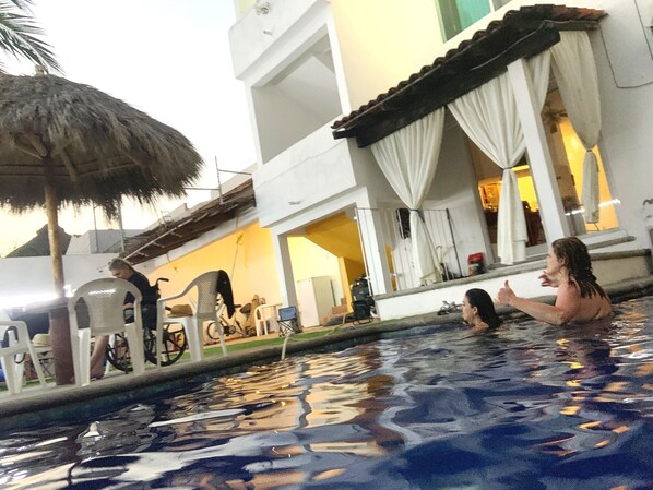 A swimming pool with a thatched roof structure, a white building with a balcony, and a person in the water.