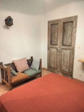 Una habitación con una cama de madera, un banco de madera con cojines, una puerta de madera y un comedero de pájaros colgado en la pared.