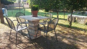 A stone table with four metal chairs, surrounded by a garden with a potted plant and a tree.
