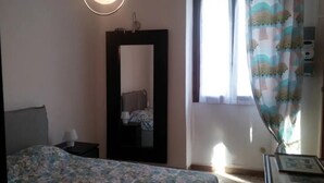 A bedroom with a bed, a nightstand, a mirror, a window with curtains, and a wall-mounted light fixture.
