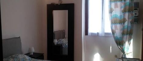 A bedroom with a bed, a nightstand, a mirror, a window with curtains, and a wall-mounted light fixture.