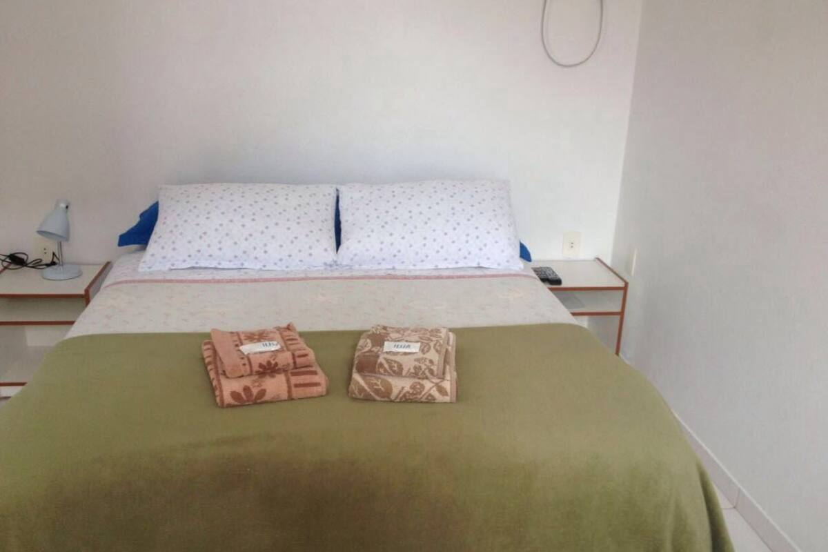 Cama bem arrumada com manta verde, duas toalhas dobradas e almofada branca estampada.