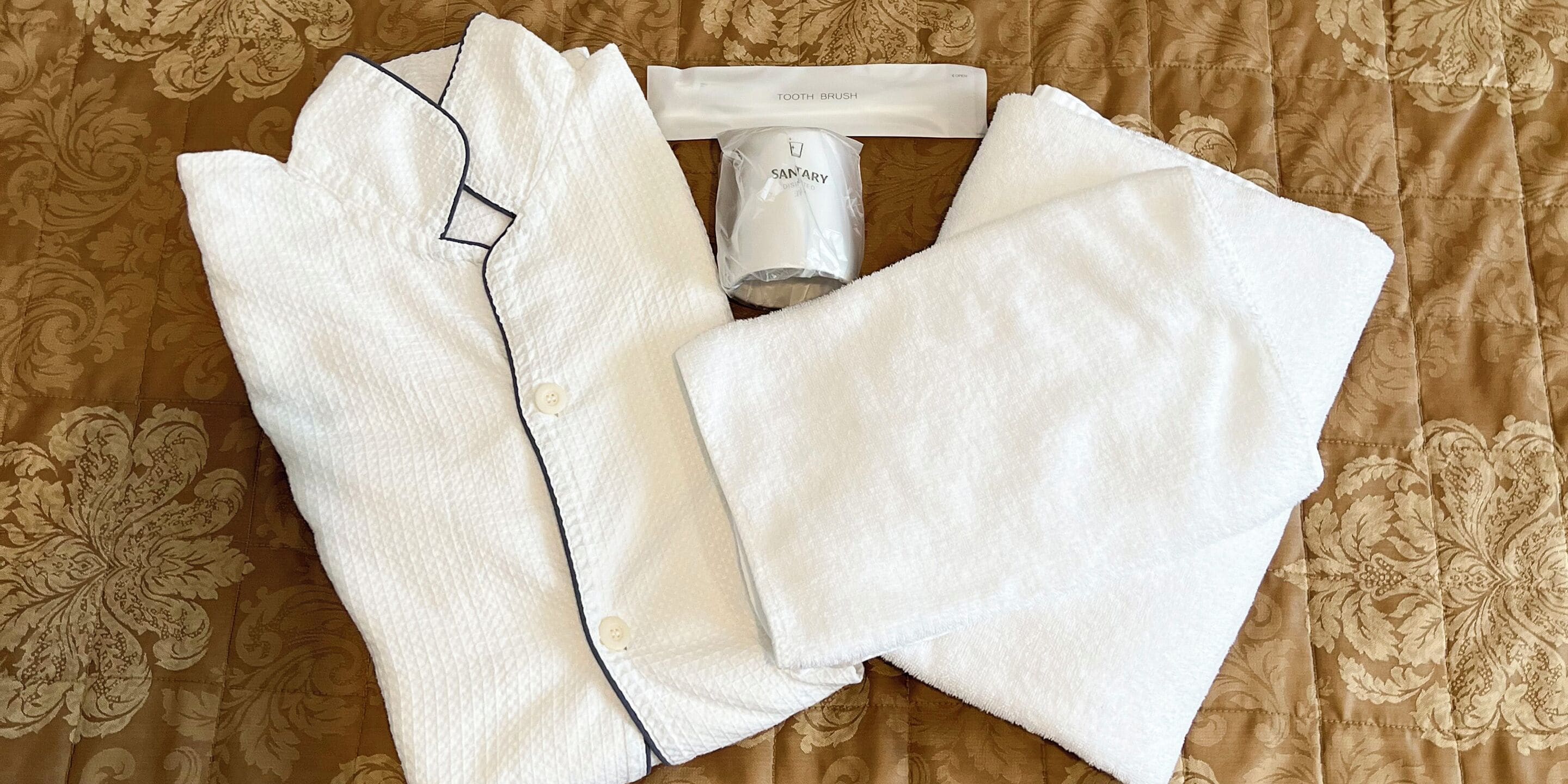 A neatly folded white shirt, a white towel, and a tube of Sanitary soap on a patterned brown bedspread.