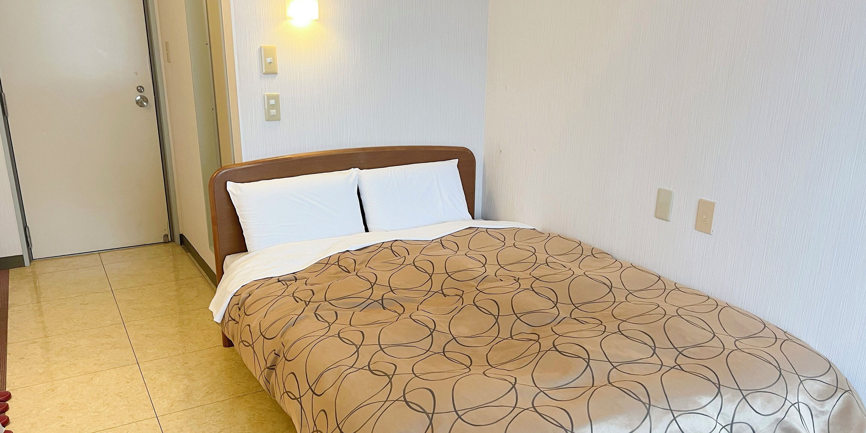 Ein Hotelzimmer mit Bett, Holz-Kopfteil, gemustertem Bettbezug und Lichtschalter an der Wand.