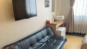 A hotel room with a black leather sofa, a flat-screen TV mounted on the wall, a desk with a telephone and a box of tissues, and a bed with a white bedspread.