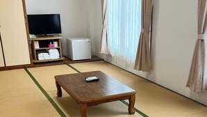 A room with tatami flooring, a wooden table, a television, a small refrigerator, and a shelf with towels. - Hotel Yamadasou (Nago)