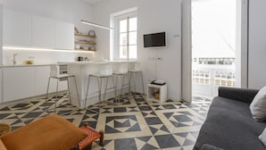 Cocina moderna con armarios blancos, una barra y una mesa de comedor. Hay un televisor montado en la pared y un balcón con barandilla.