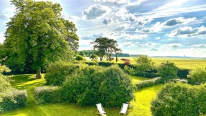 Ein gepflegter Garten mit geschnittenen Hecken, zwei leeren Liegestühlen und einem großen Baum, der Schatten spendet.