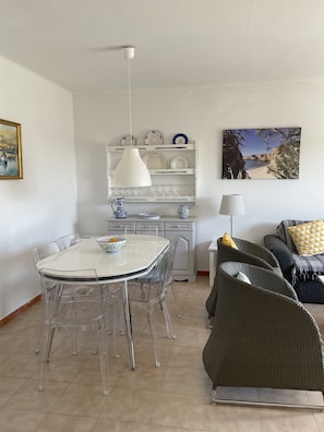 A modern living room with a dining area, featuring a white table, clear chairs, wicker furniture, and a coastal painting.