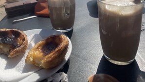A table with pastries and two glasses of iced coffee, overlooking a beach with people in the background.