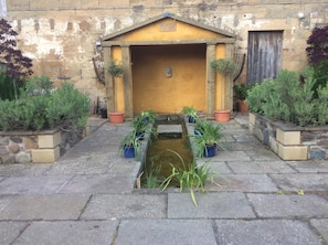 Un patio con un pequeño elemento acuático, plantas en macetas y un edificio de fachada amarilla.