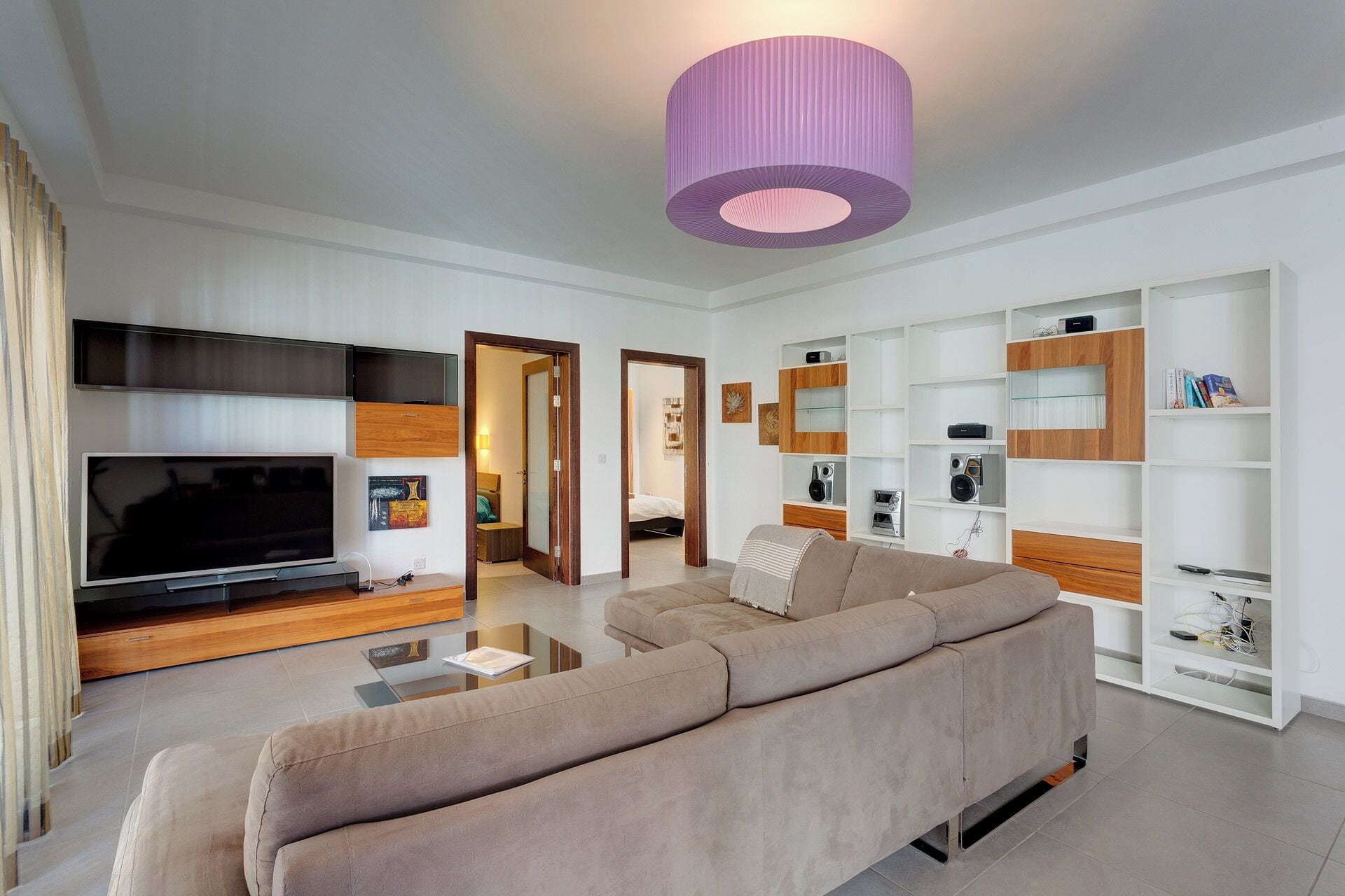 Un salon moderne avec un grand téléviseur à écran plat, un canapé beige et une table basse en verre. On y trouve des étagères en bois où sont disposés divers objets, ainsi qu’un luminaire cylindrique violet suspendu au plafond.