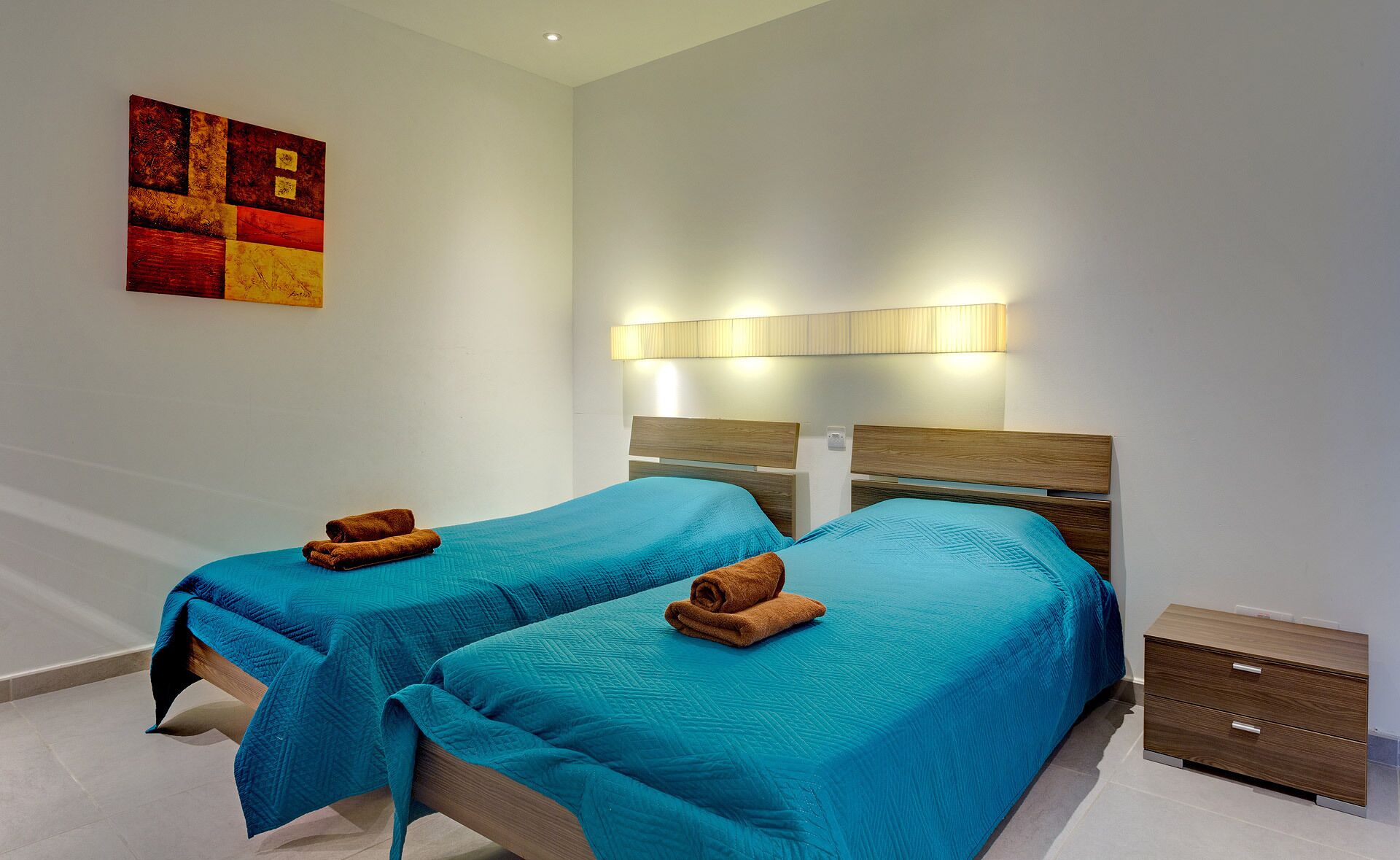 A hotel room with two single beds, each with a teal bedspread and a brown towel. There is a wall-mounted light fixture above the beds and a colorful abstract painting on the wall.