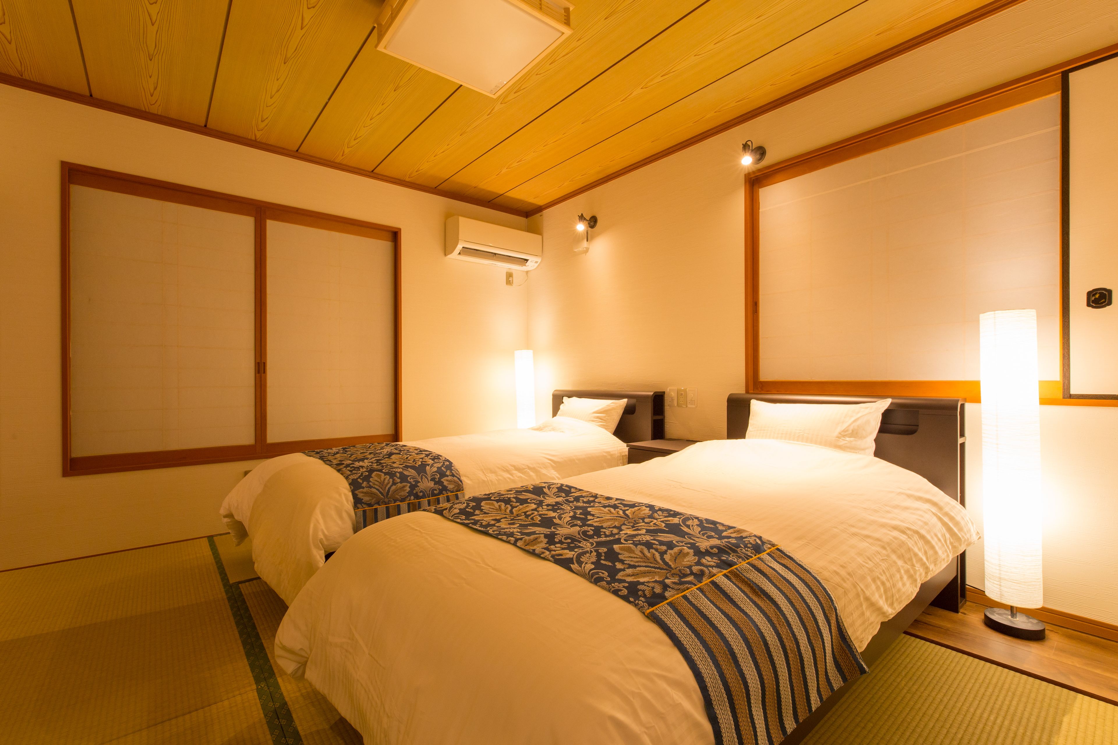 A hotel room with two beds, a wooden ceiling, an air conditioning unit, and a bedside lamp.