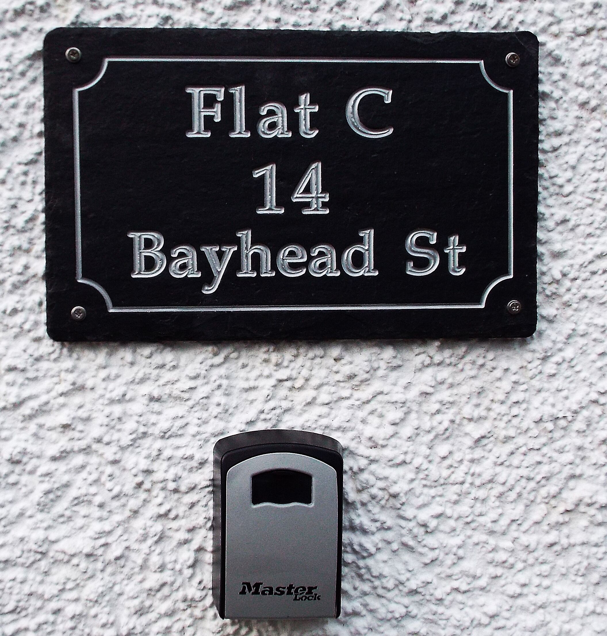 A black metal sign with white lettering indicating 'Flat C, 14 Bayhead St.' Below the sign, there is a metal door lock labeled 'Master Lock.'