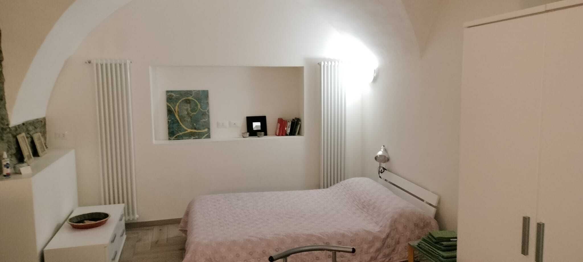 Ein Schlafzimmer mit einem Bett, einem Nachttisch, einem Heizkörper, einem Regal mit Büchern und einem Bild sowie einem Kleiderschrank.