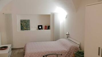 Ein Schlafzimmer mit einem Bett, einem Nachttisch, einem Heizkörper, einem Regal mit Büchern und einem Bild sowie einem Kleiderschrank.