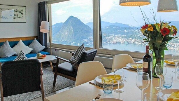 A hotel room with a large window offering a view of a mountain and city. There is a dining table set with plates, glasses, and a bottle of wine. The room has a sofa with pillows and a chair. - Nice apartment in villa with WIFI, private pool, TV, terrace, pets allowed and panoramic view (Lugano)