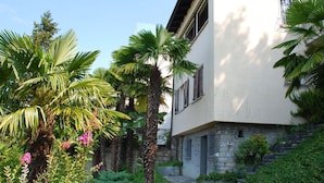 A two-story building with a stone base and white upper walls, surrounded by lush greenery and palm trees. - Nice apartment in villa with WIFI, private pool, TV, terrace, pets allowed and panoramic view (Lugano)