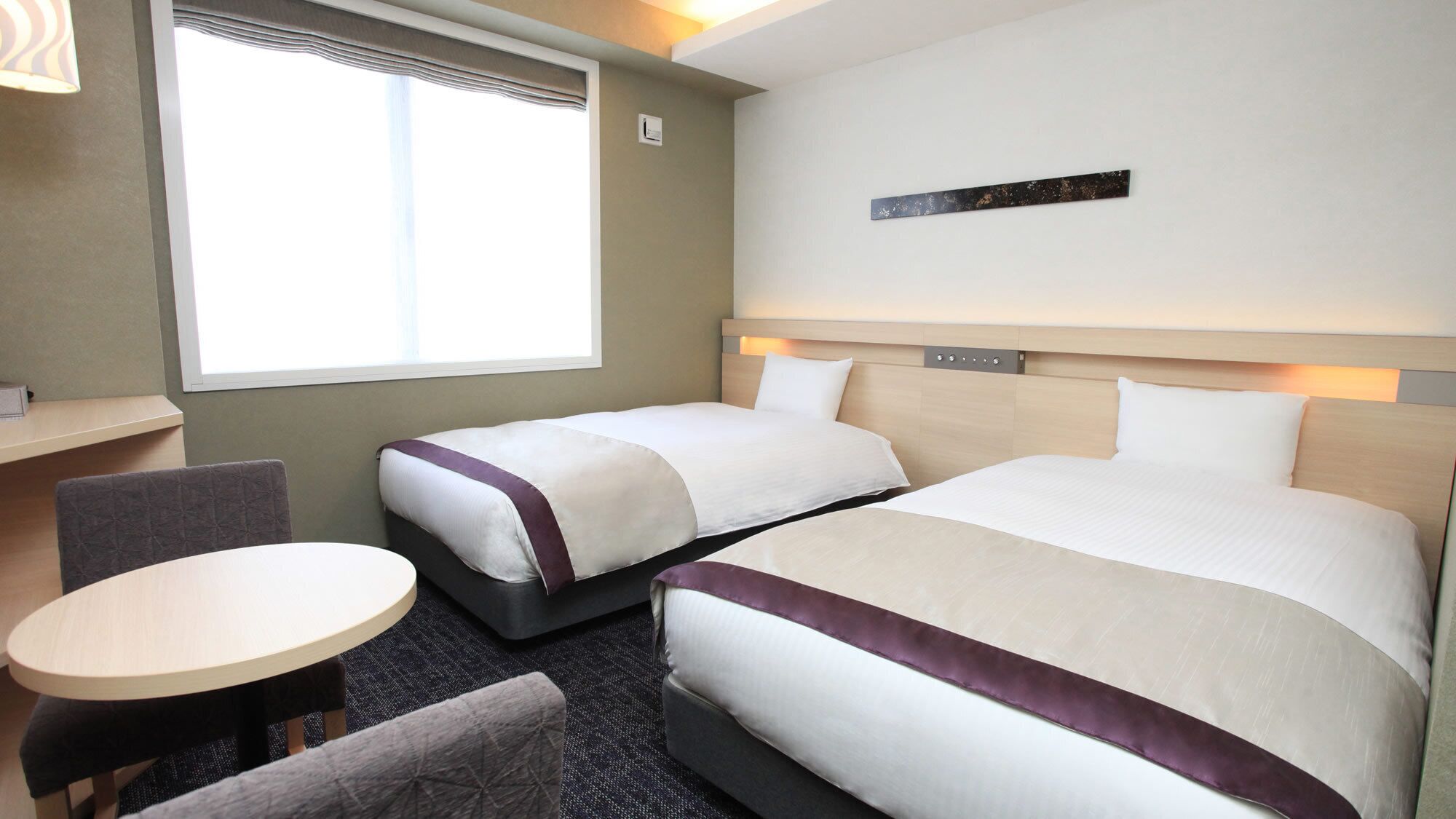 Quarto de hotel com duas camas, uma mesinha, uma cadeira e uma janela com persianas.