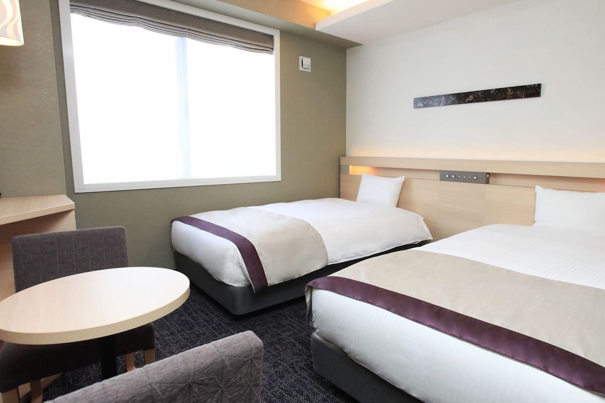 Quarto de hotel com duas camas, uma mesinha, uma cadeira e uma janela com persianas.