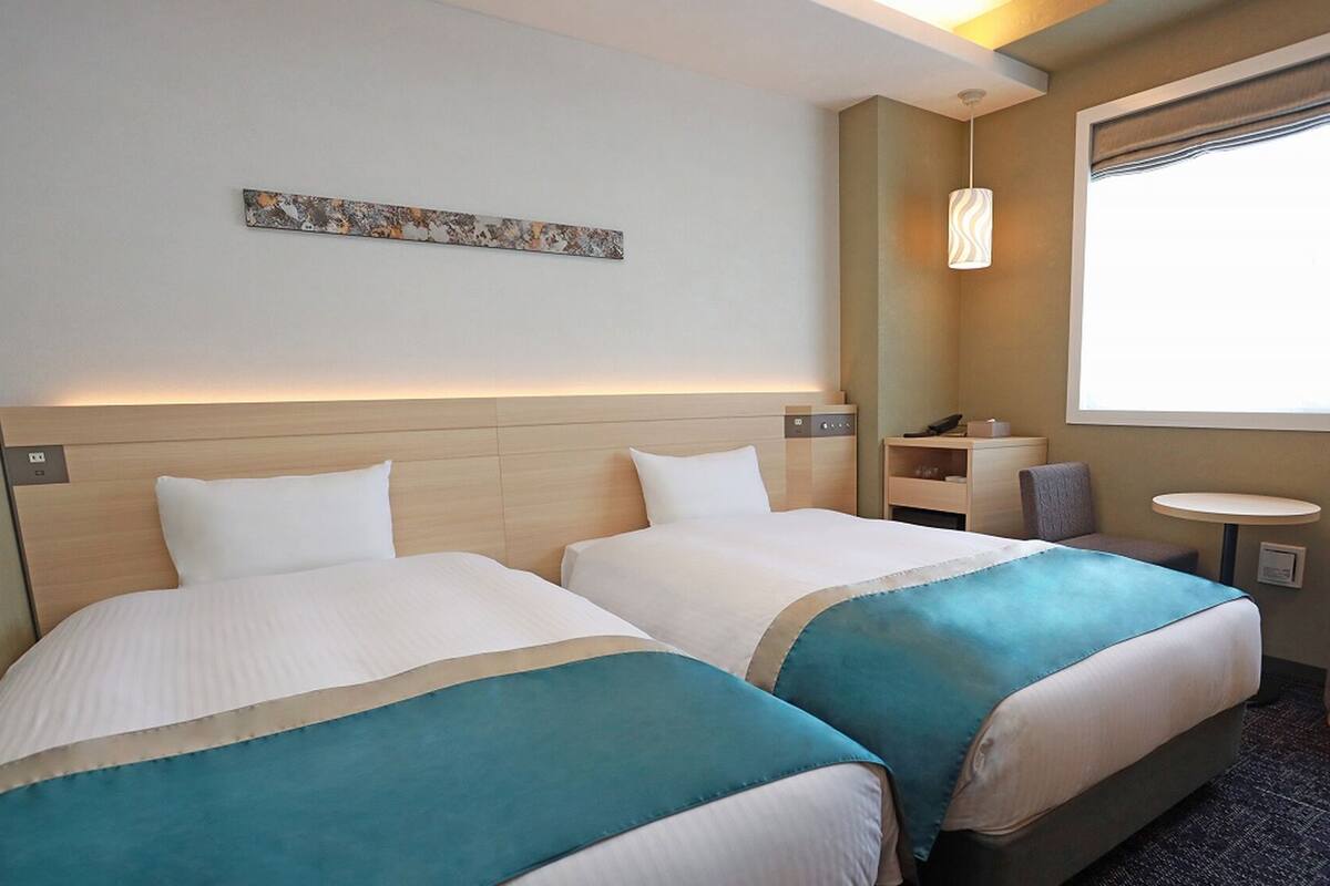 A hotel room with two beds, a small table, a chair, a lamp, and a window with a view.