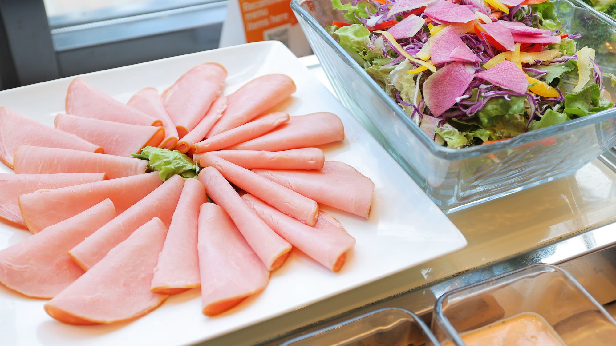 A buffet table with a plate of sliced ham arranged in a circular pattern and a glass bowl containing a salad with various vegetables and radish slices.