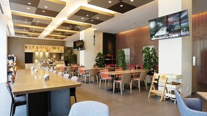 A modern restaurant with a long communal table, individual tables, and chairs. There are potted plants and a television mounted on the wall. - Urban Hotel Kyoto Shijo Premium (Kyoto)