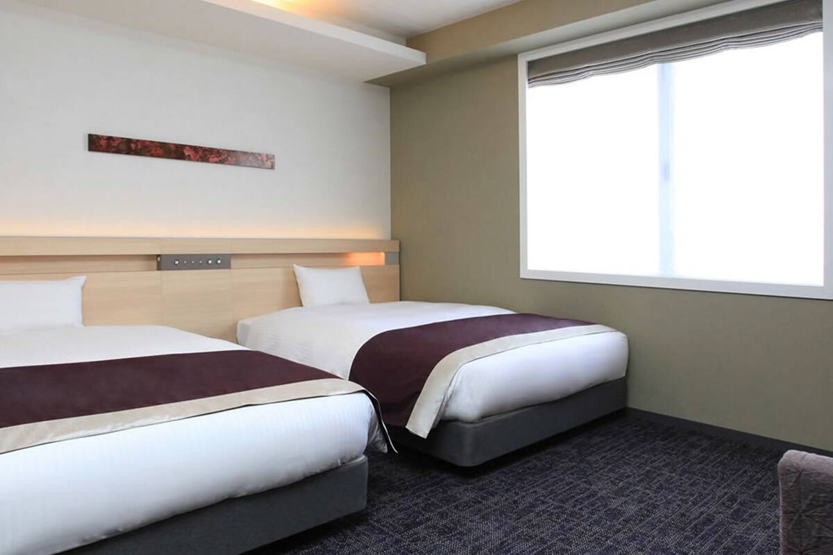 Quarto de hotel com duas camas, janela com persianas, cabeceira com painel de luz e uma pequena obra de arte na parede.