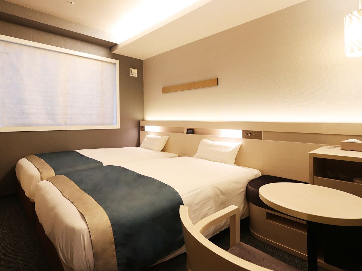 Habitación de hotel con dos camas, un escritorio, una silla, una ventana con persianas y un estante en la pared.