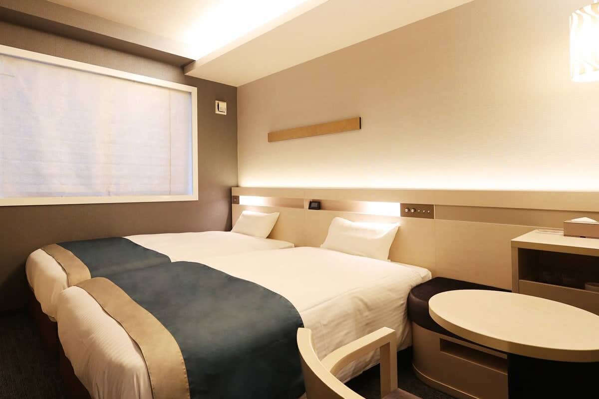 Habitación de hotel con dos camas, un escritorio, una silla, una ventana con persianas y un estante en la pared.