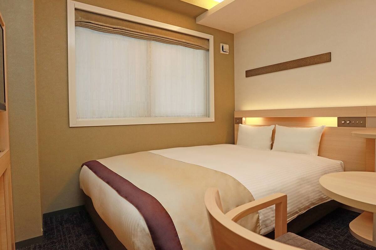 A hotel room with a bed, bedside table, chair, and a small table with a lamp.