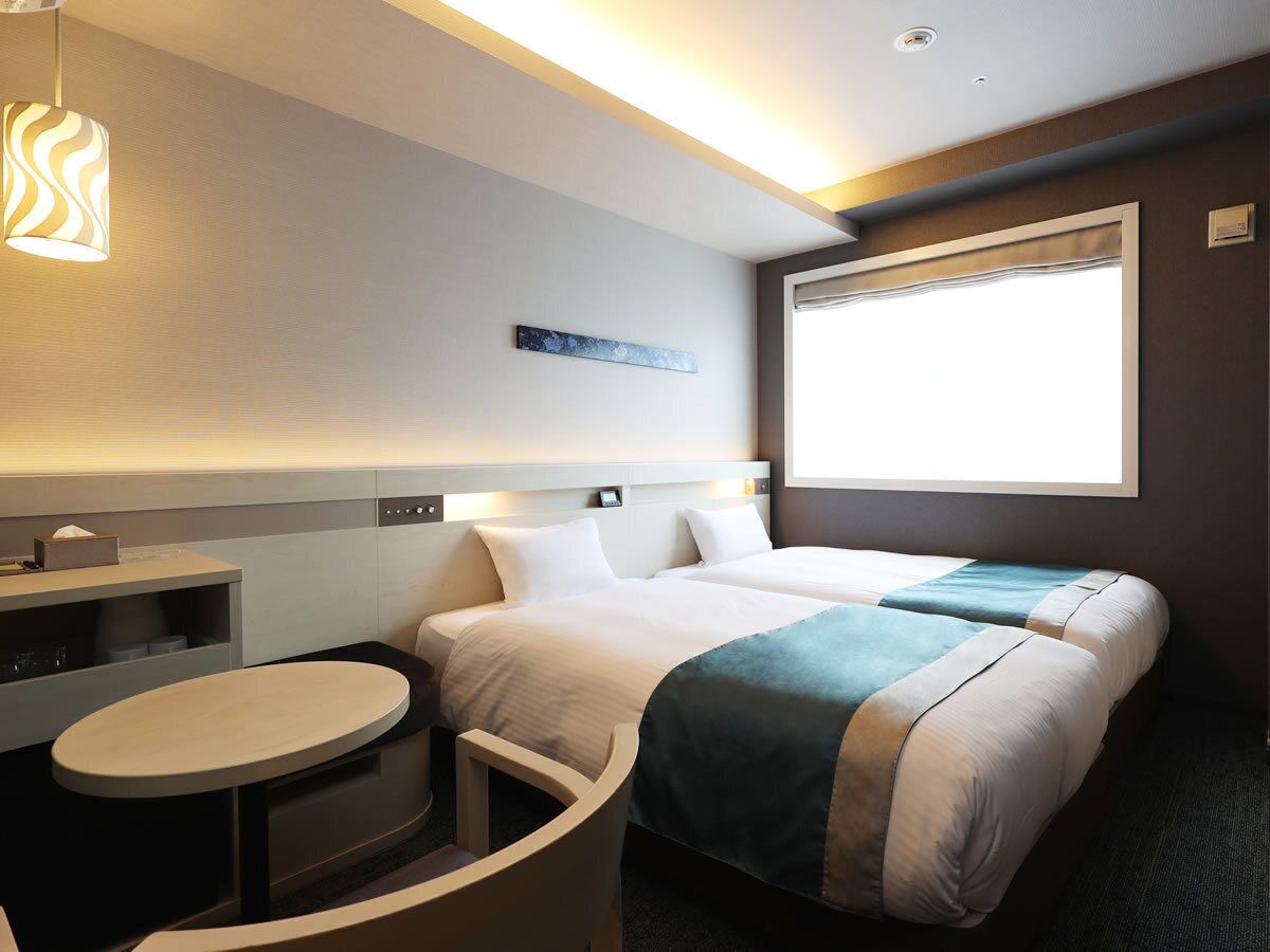 Quarto de hotel com duas camas, uma mesa pequena, prateleira na parede e janela com vista.