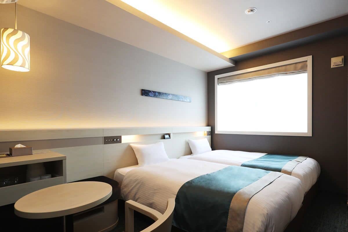 Quarto de hotel com duas camas, uma mesa pequena, prateleira na parede e janela com vista.