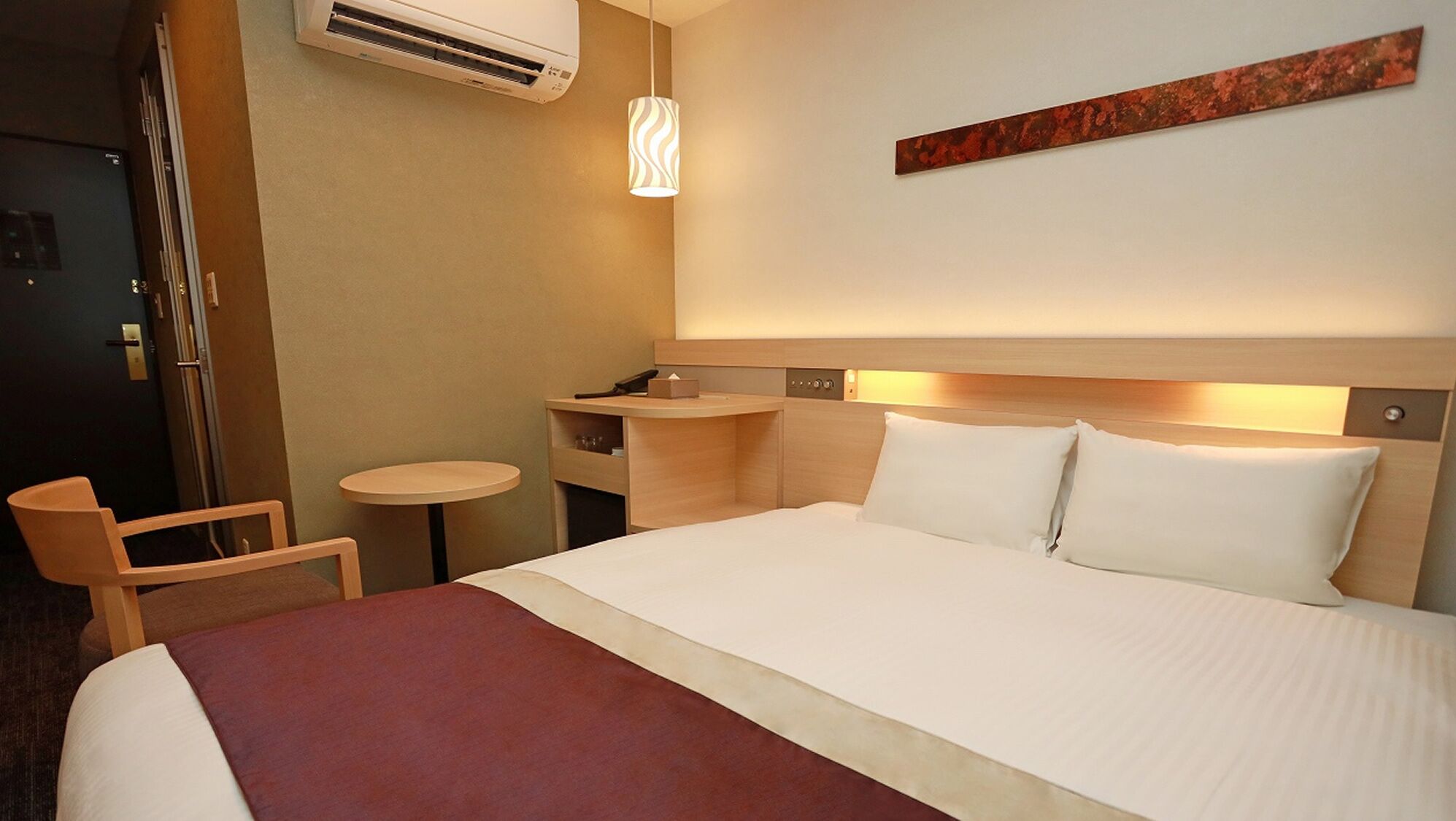 Habitación de hotel con cama, mesita de noche, silla, escritorio y unidad de aire acondicionado.