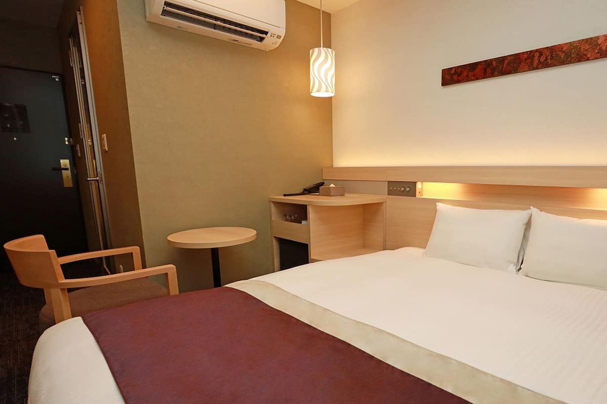 Habitación de hotel con cama, mesita de noche, silla, escritorio y unidad de aire acondicionado.