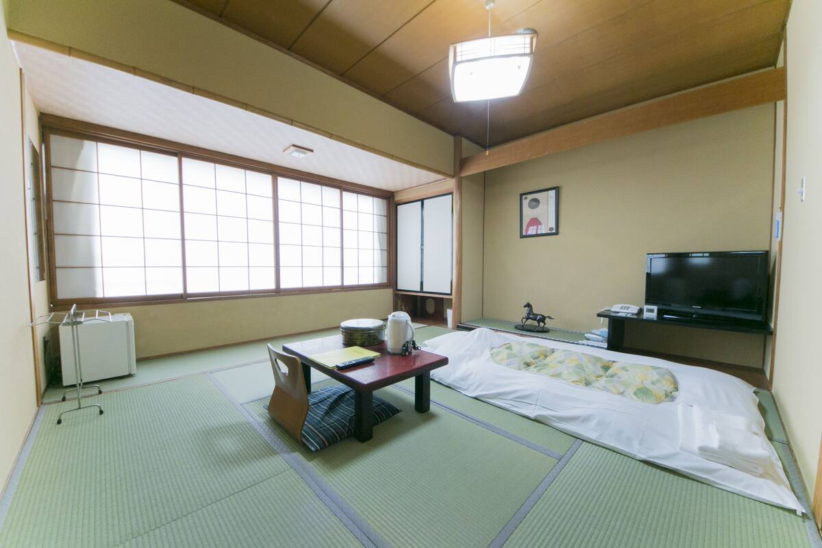 Ein traditioneller japanischer Raum mit Tatami-Böden, einem Futon, einem Fernseher und einem kleinen Tisch mit Wasserkocher und Tassen.