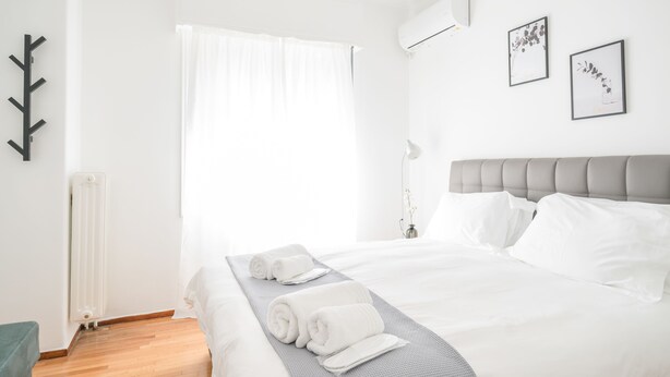 A neatly made bed with white linens and rolled towels, a grey headboard, and a radiator on the wall. - Modern Design Apartment Near Acropolis 2 (Athens)