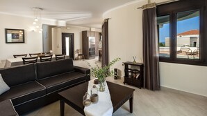 A modern living room with a dark brown sofa, a coffee table with decorative items, and a view of the outdoor terrace through large windows. - Villa Stellina, elegant villa, full privacy (Chania)