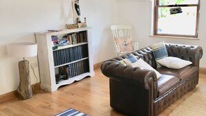 A cozy living room with a brown leather sofa, a white bookshelf, a striped rug, and a window with a view of the sea.