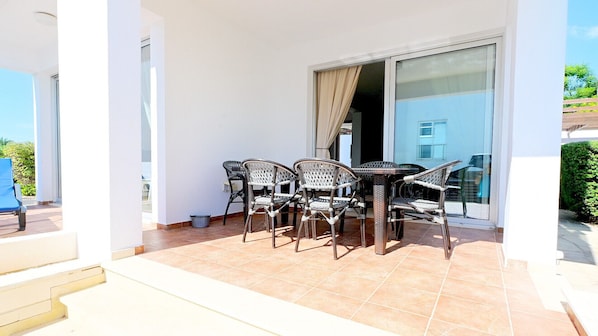 A covered outdoor seating area with a table and chairs, a sliding glass door, and a view of the surrounding landscape.