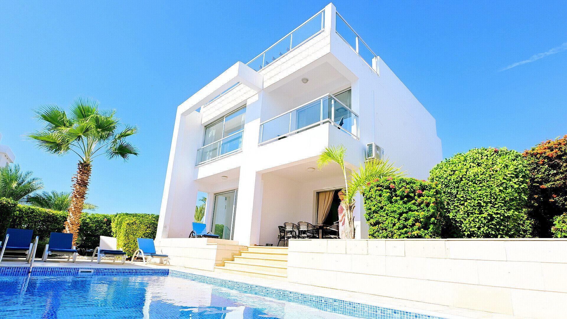 A modern two-story building with a swimming pool, outdoor seating, and palm trees.