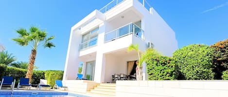 A modern two-story building with a swimming pool, outdoor seating, and palm trees.