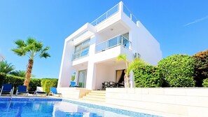 A modern two-story building with a swimming pool, palm trees, and a well-maintained garden.