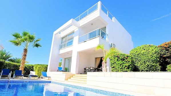 A modern two-story building with a swimming pool, outdoor seating, and palm trees.