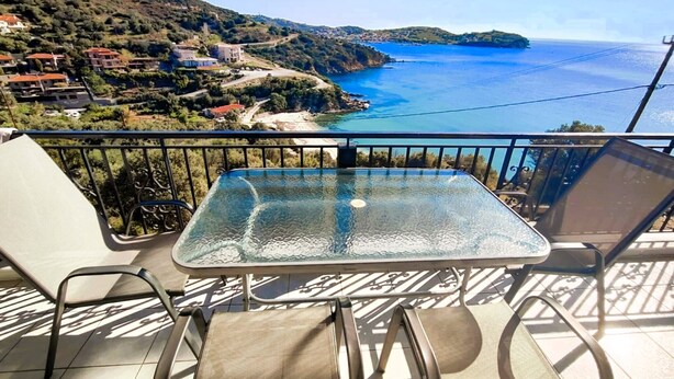 Un balcón con una mesa de vidrio y dos sillas, con vistas a un paisaje costero con casas y un mar azul cristalino.